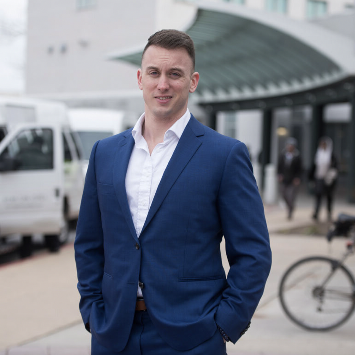 Photo of a young man in a blue suit smiling in front of a busy sidewalk